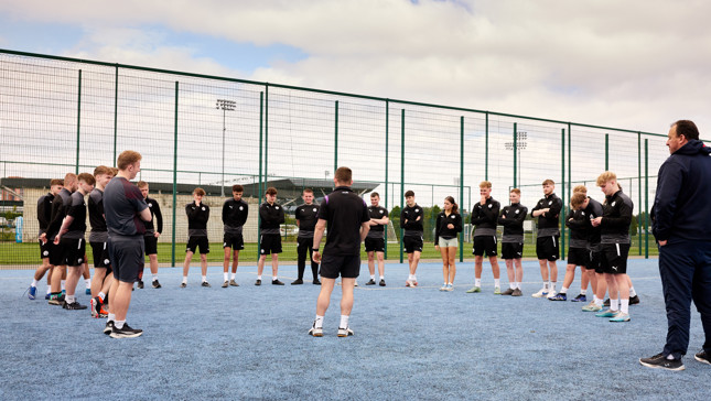 A group of young athletes huddled in a circle on a blue outdoor sports court, wearing sports uniforms, likely receiving instruction or motivation from a coach standing in the center.