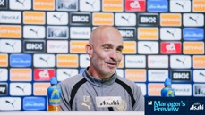 A man in a Manchester City jacket speaks at a press conference. The background displays various sponsor logos including Puma and Etihad.