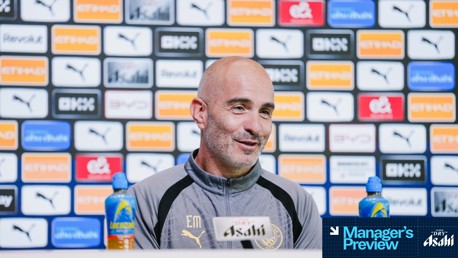 A man in a Manchester City jacket speaks at a press conference. The background displays various sponsor logos including Puma and Etihad.