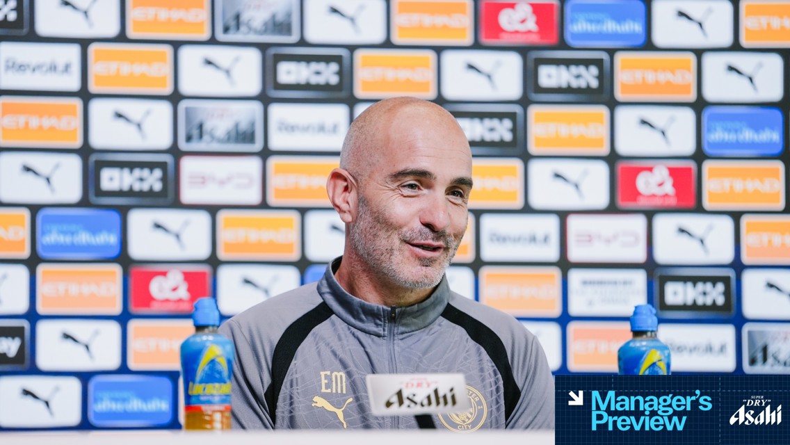 A man in a Manchester City jacket speaks at a press conference. The background displays various sponsor logos including Puma and Etihad.