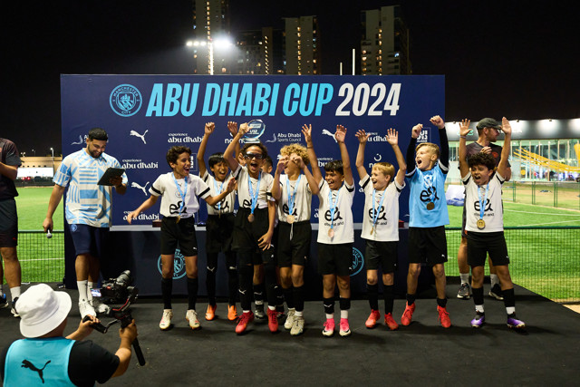 A group of young football players celebrate on stage after their victory at the Abu Dhabi Cup 2024. The team is wearing medals and standing in front of an event backdrop. Coaches and a videographer are also visible.