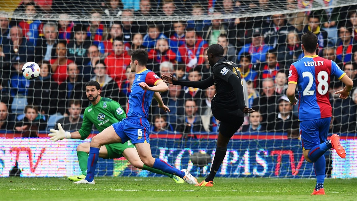 A football match scene where a player in a black uniform attempts to score a goal against a team in blue and red uniforms. The goalkeeper is in a green uniform reaching out to block the ball.