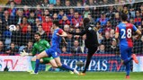 A football match scene where a player in a black uniform attempts to score a goal against a team in blue and red uniforms. The goalkeeper is in a green uniform reaching out to block the ball.