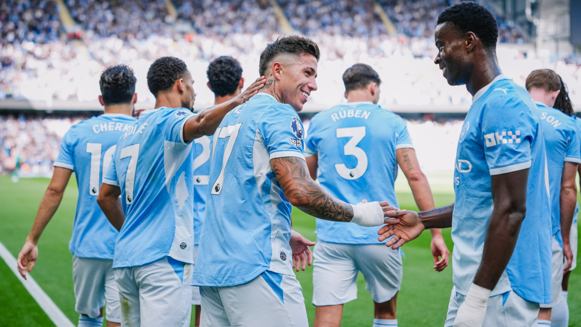 Group of Manchester City players in blue football kits celebrating on the field, with visible names and numbers on their backs.