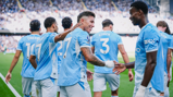 Group of Manchester City players in blue football kits celebrating on the field, with visible names and numbers on their backs.