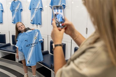 Two fans are taking a selfie holding a Manchester City jersey with the name 'Grealish' and number 10 at the Manchester City dressing room. Other jerseys are hanging in the background.