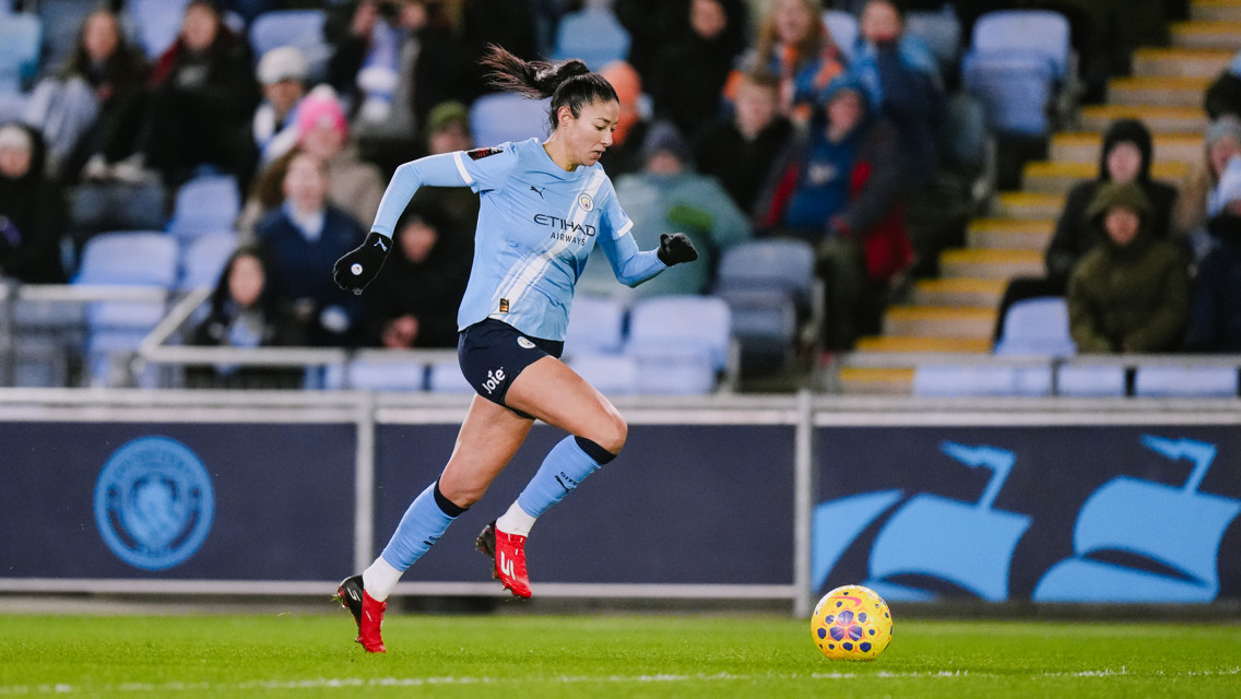 A woman soccer player in a Manchester City uniform is running on a field during a match. She is wearing red cleats and a black glove. The stadium crowd is visible in the background.