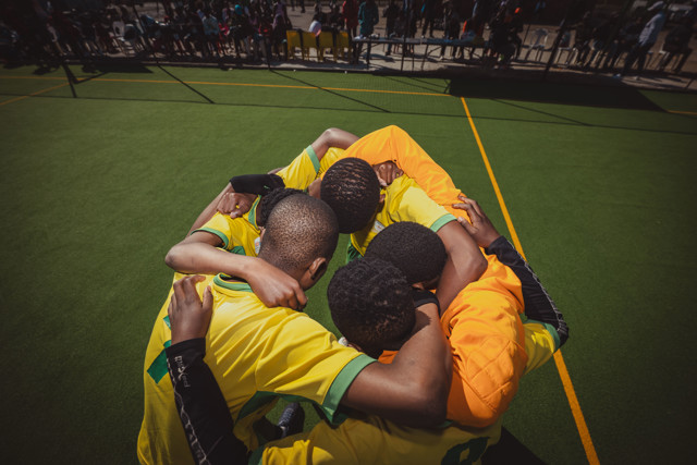 A group of soccer players in yellow and orange jerseys huddle together on a green pitch with spectators in the background.