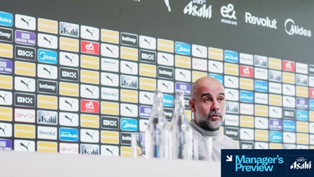 Press conference backdrop featuring multiple sponsor logos, water bottles, and partial view of a person at the table.