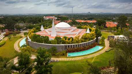 Aerial view of a modern architectural building with conical structures, surrounded by landscaped gardens and water features, set against a backdrop of greenery and distant hills.