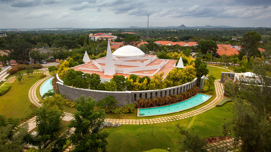 Aerial view of a modern architectural building with conical structures, surrounded by landscaped gardens and water features, set against a backdrop of greenery and distant hills.