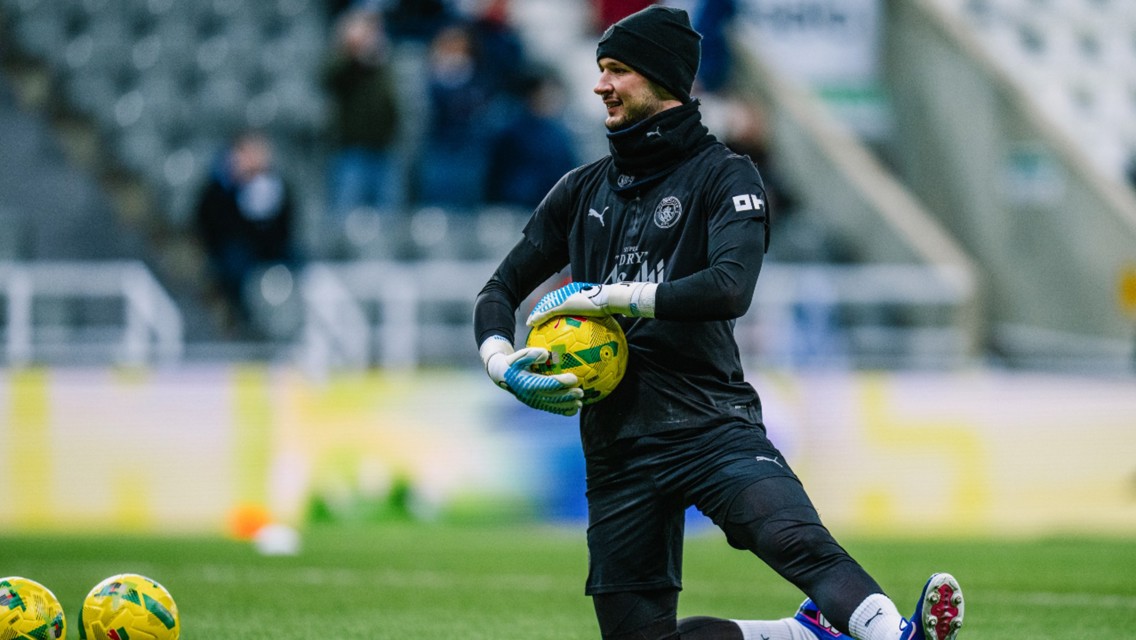 A soccer goalkeeper wearing black training gear and gloves kneeling on the field with a yellow ball in hand. Multiple yellow soccer balls are visible on the grass nearby.