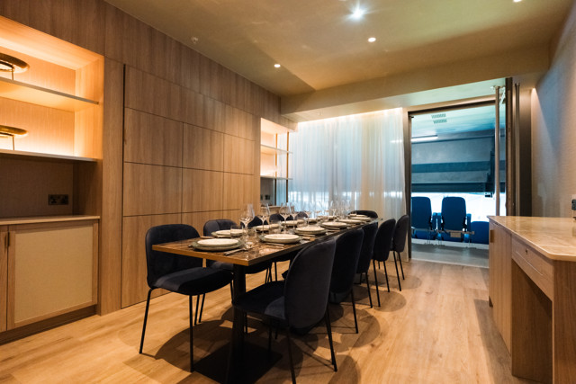A modern dining room with a long table set for dinner, featuring elegant lighting and wooden panels. There is a glimpse of a private viewing area with blue chairs, separated by translucent curtains.
