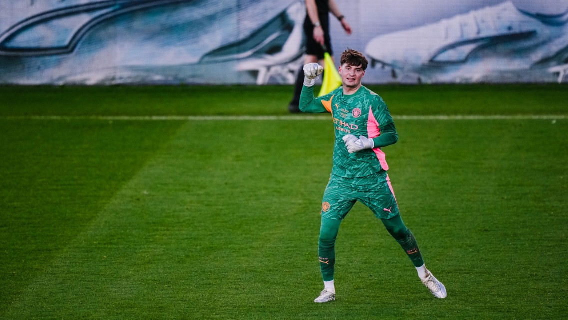 A football goalkeeper wearing a green and pink kit with Etihad Airways branding, on a grass field, making a fist gesture.
