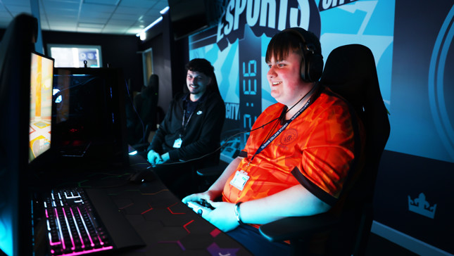 Two people are sitting at gaming stations in an esports environment. One wears a red shirt with a name badge, and both have headphones and blurred faces.