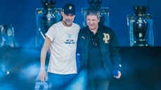 Two individuals on stage with Manchester City logos in the background and trophies displayed. One wears a white t-shirt with text, and both have caps and jackets with 'P' logos.
