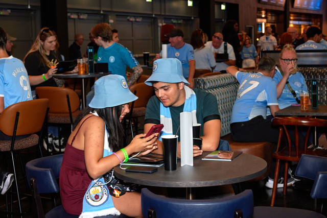 Fans in Manchester City jerseys and hats are socializing and checking a phone in a club setting, with other fans in the background.