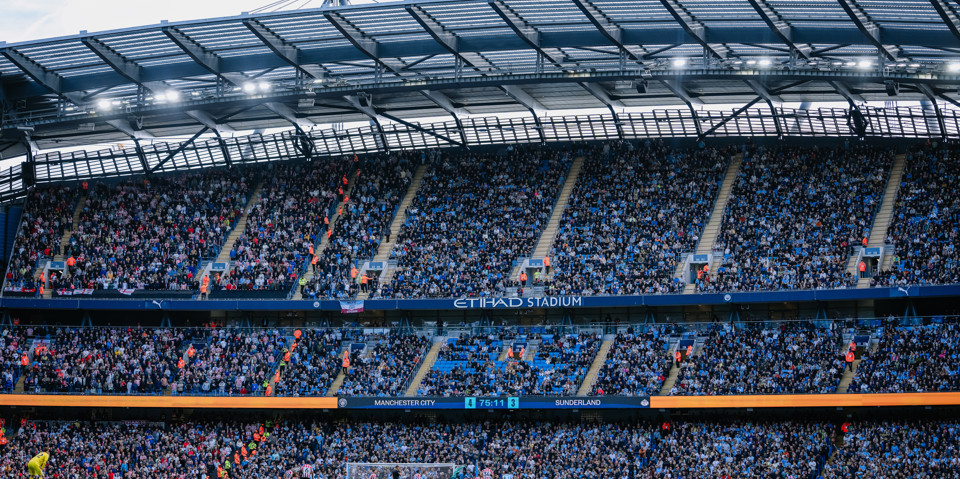 A packed Etihad Stadium during a football match between Manchester City and Sunderland. The score is displayed as 4-3, and the crowd fills the stands.