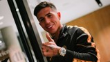 Person in a black and gold tracksuit giving a thumbs-up gesture. They have tattoos on their neck and hand and are wearing a watch with a white face.