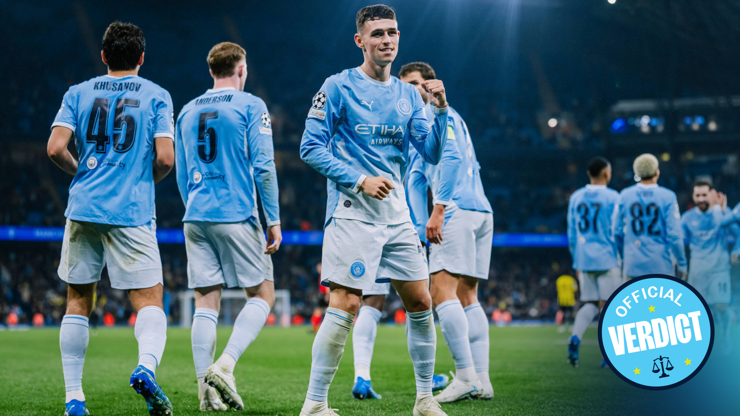 Players in Manchester City jerseys celebrating a goal on a soccer field during a match with a stadium crowd in the background.