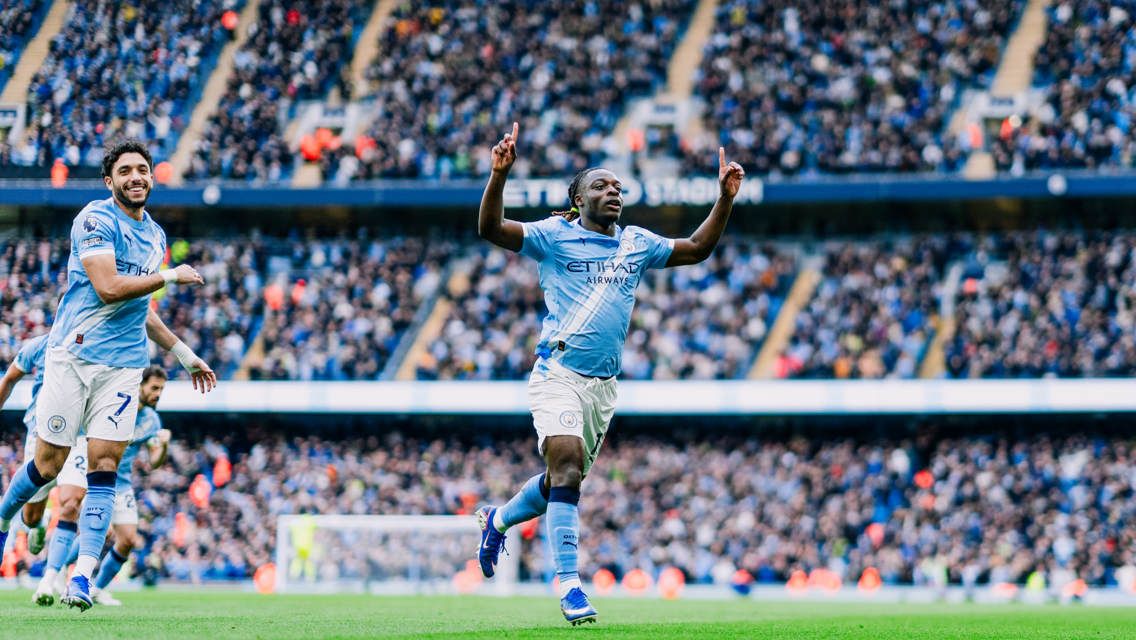 Soccer players in light blue Manchester City uniforms celebrating a goal during a match at a crowded stadium.