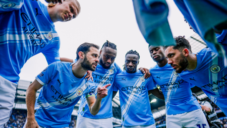 A group of Manchester City football players in a huddle, wearing blue jerseys, during a team talk on the field.