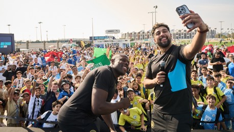 A large crowd gathers for a selfie with two men on stage, one holding a microphone. People in the background wave and wear various colored shirts and hold flags.