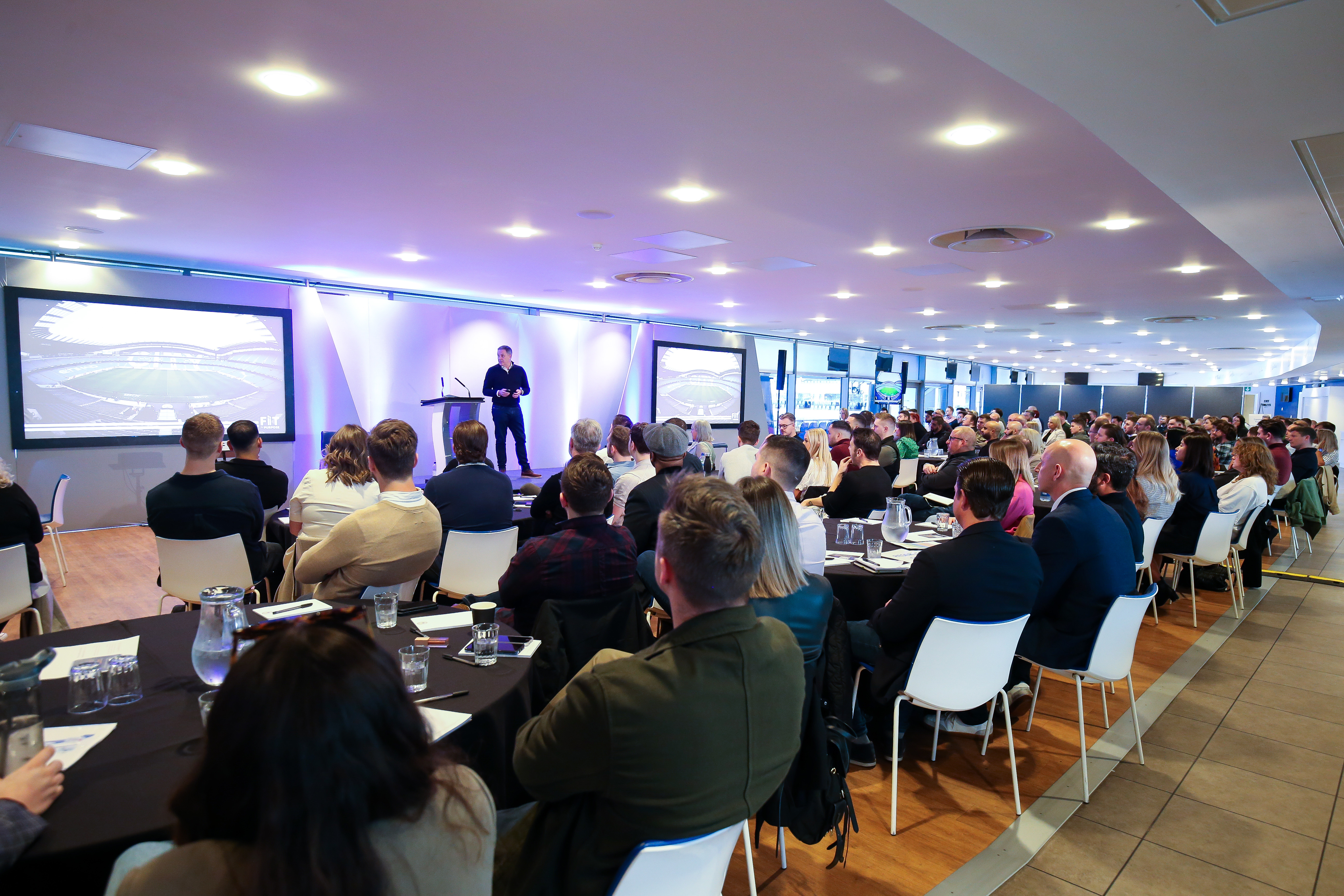 A speaker presents to a large audience in a modern conference room with two large screens displaying a stadium image.
