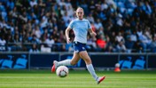 A football player in a light blue jersey and shorts runs on the field during a match, with a crowd in the background.