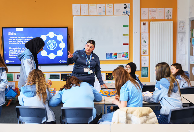 A classroom scene with students and a teacher conducting a workshop. The digital screen displays educational content related to Manchester City skills. Students are engaged in learning activities around a table.