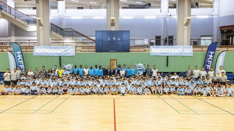 Large group of children and adults in a sports hall posing together for a photograph. They are wearing blue Manchester City shirts with mascots and banners in the background.