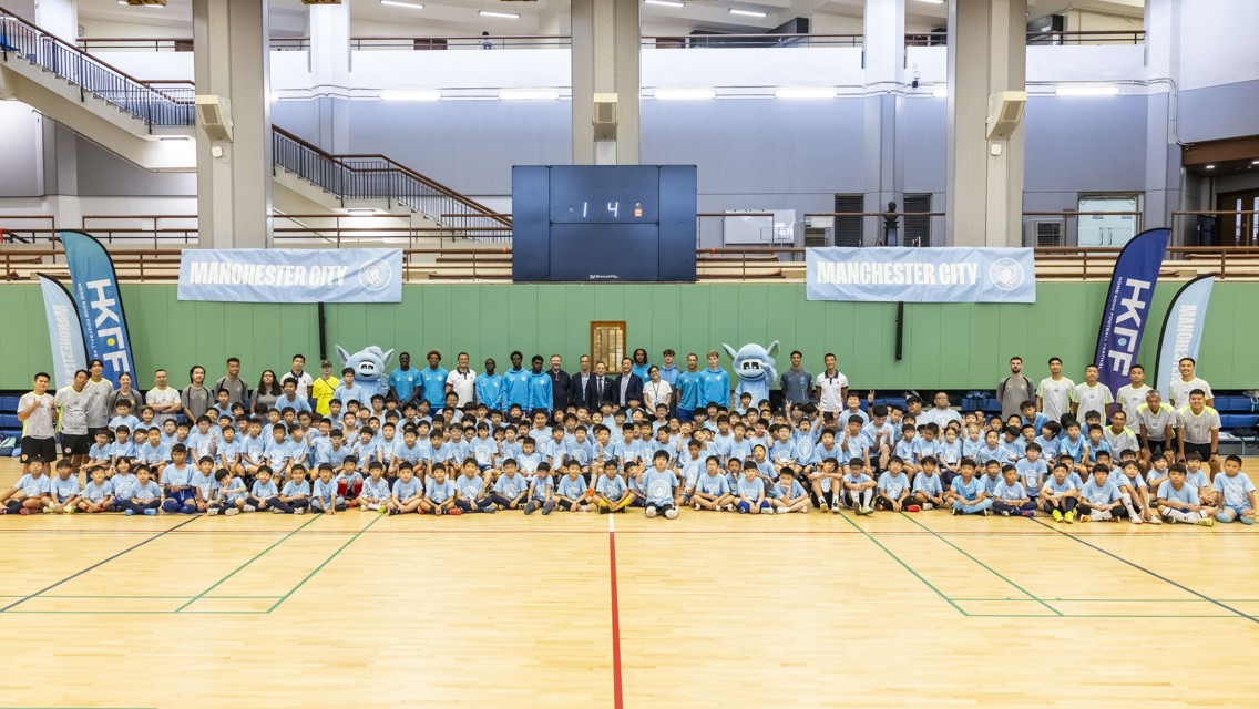 Large group of children and adults in a sports hall posing together for a photograph. They are wearing blue Manchester City shirts with mascots and banners in the background.