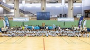 Large group of children and adults in a sports hall posing together for a photograph. They are wearing blue Manchester City shirts with mascots and banners in the background.
