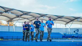 A group of people in a military-style fitness training exercise wearing camouflage pants and t-shirts indoors at a stadium with blue seating and a covered canopy.