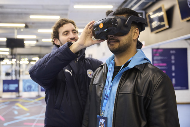 Two people interact with a VR headset at a stadium, featuring Manchester City logos and Puma branding.