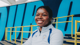 A smiling woman in a Manchester City tracksuit sitting on stadium steps with a vibrant blue and yellow background.