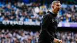 A coach wearing Manchester City's crest stands near Etihad Stadium with a crowd in the background.