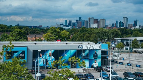Exterior view of the Manchester City 'City Store' with large player images on display and cars parked in front, city skyline in the background.