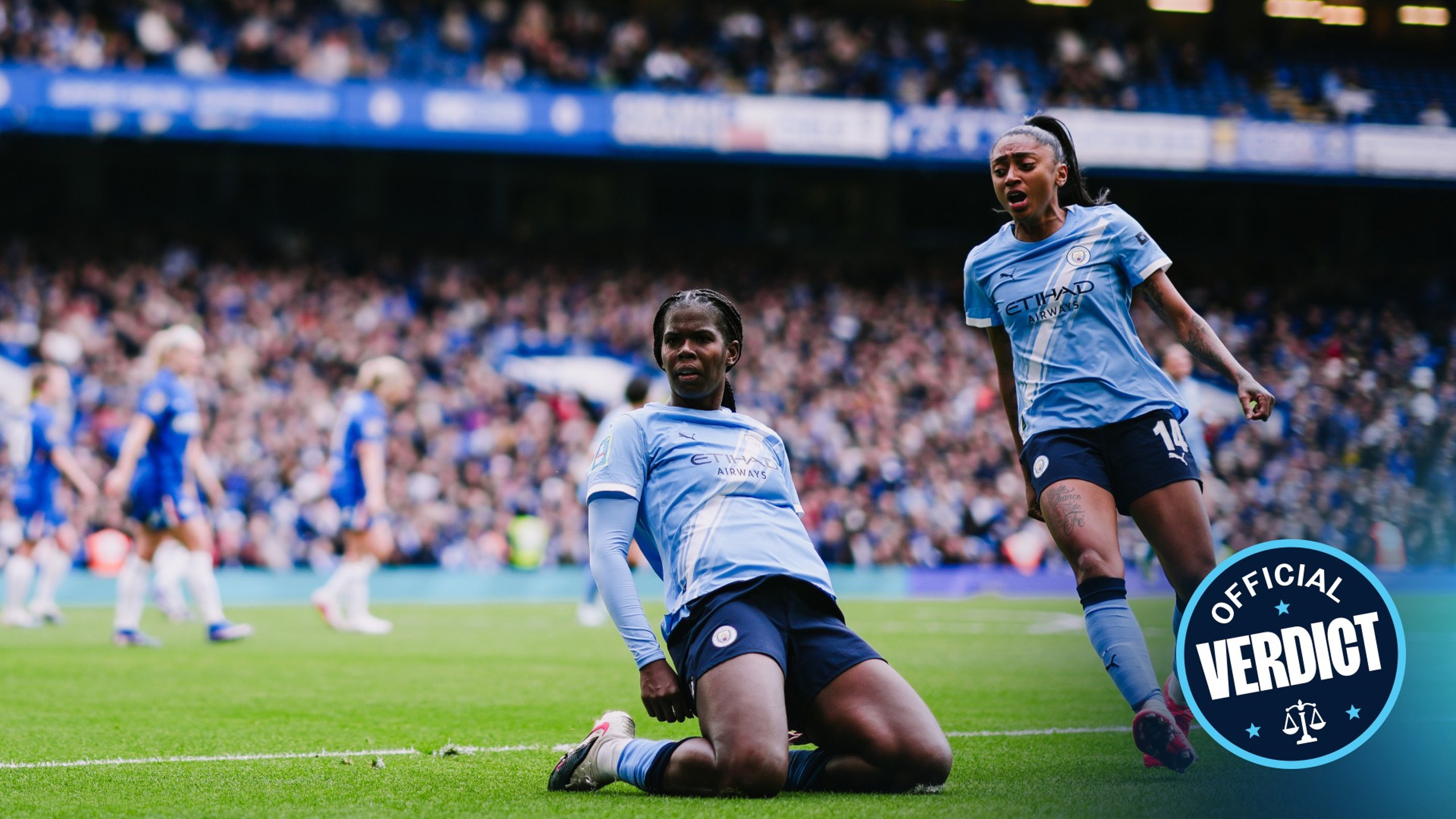 Two football players from Manchester City Women's Team celebrate after scoring a goal against Chelsea. The players are wearing blue Manchester City kits. The scene is set in a stadium with blurred spectators in the background.