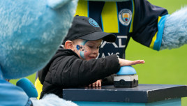 A child wearing Manchester City merchandise presses a button at a mascot event, accompanied by a Manchester City mascot.