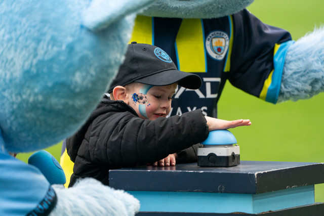 A child wearing Manchester City merchandise presses a button at a mascot event, accompanied by a Manchester City mascot.