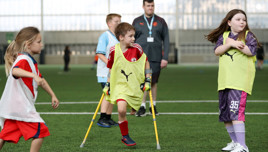 Children playing adaptive football on a field indoors, one child using crutches, all wearing Puma bibs.
