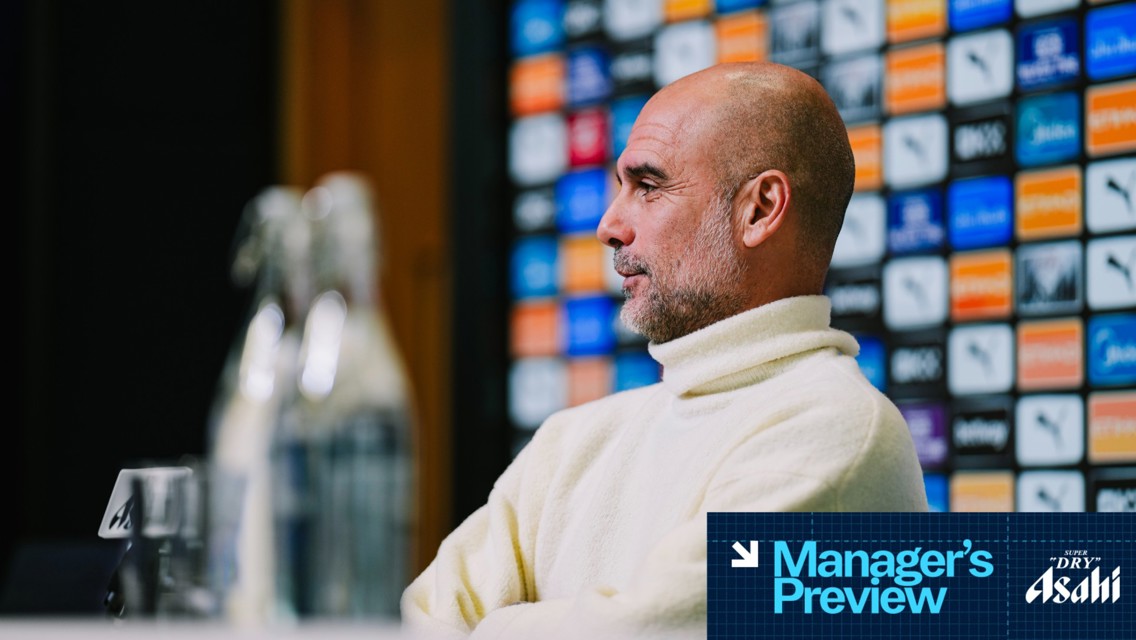 A person with blurred face at a press conference wearing a white sweater, seated next to branded bottles and a wall covered in media sponsor logos.