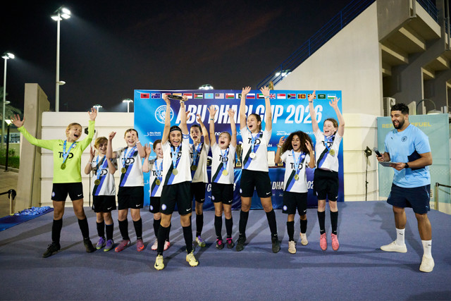 A youth soccer team celebrates on stage after winning at the Abu Dhabi Cup 2026. They wear medals and hold trophies, with a banner behind them displaying flags and event details.