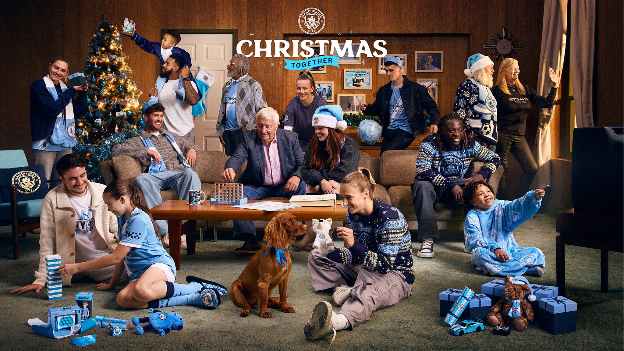 A Manchester City-themed Christmas gathering with people wearing blue jerseys, a decorated tree, gifts, mugs, a dog and toy bear, set in a cozy living room.