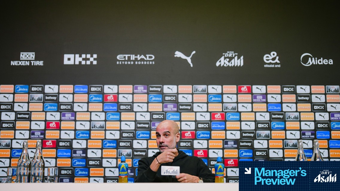 Manchester City press conference backdrop featuring multiple sponsor logos. The blurred individual at the podium has bottles on the table along with branded drink containers.