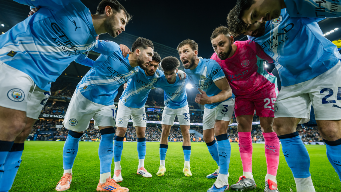 Manchester City players in a team huddle wearing blue and white kits on a football field at Etihad Stadium.