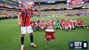 A Norwegian soccer player wearing a red jersey with the number 9 is about to hit a red drum with a drumstick. Behind, a group of soccer teammates in matching jerseys sit on the grass celebrating. The event occurs in a large stadium filled with spectators.