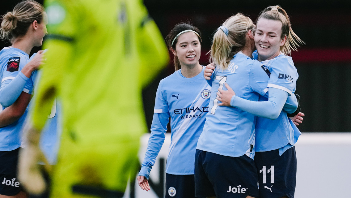 Manchester City Women's football players celebrating during a match. The players are wearing light blue kits, featuring the Manchester City crest and sponsor logos.