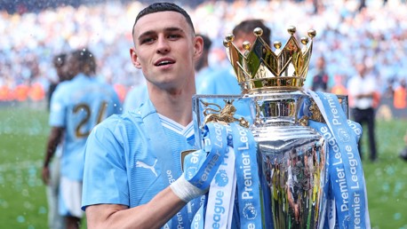 A person in a Manchester City jersey holds the Premier League trophy. The crowd and players in similar jerseys are in the background.